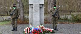 Na środku kamienny pomnik w kształcie krzyża. Po jego prawej i lewej stronie stoi żołnierz. Pod pomnikiem leżą złożone wieńce i stoją zapalone znicze