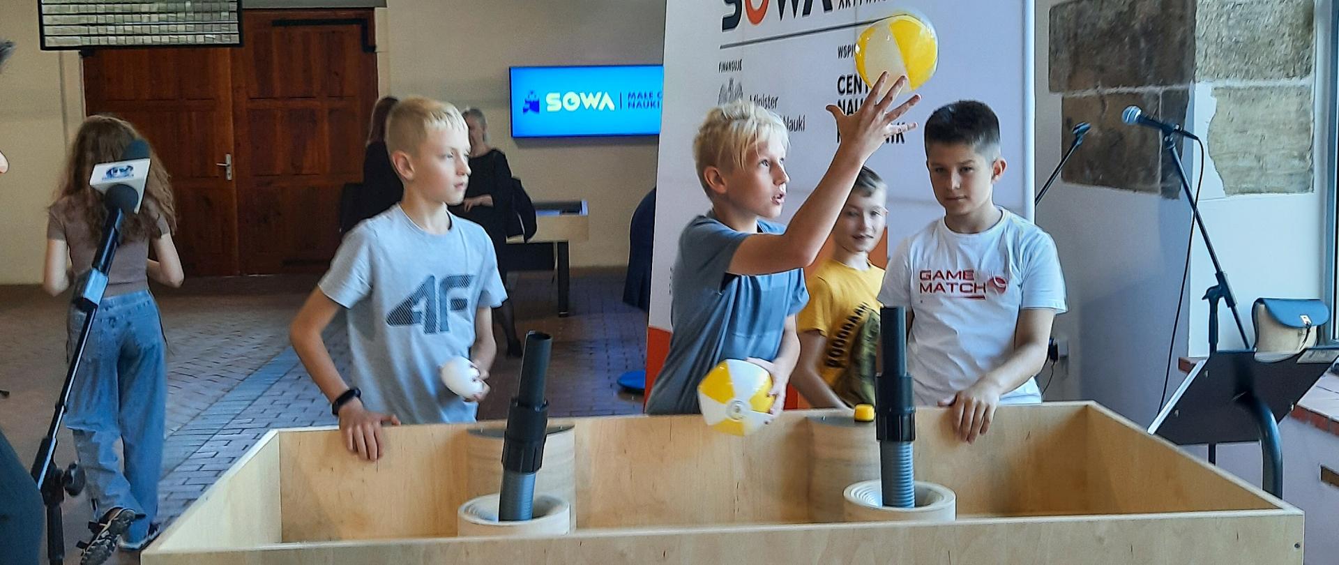 Grupa dzieci podrzuca żółte piłki, które unoszą się nad drewnianą skrzynią, za nimi baner z napisem SOWA.