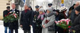 Kilkanaście osób stoi przed bazyliką, trzy trzymają w ręku wiązanki kwiatów, pada śnieg