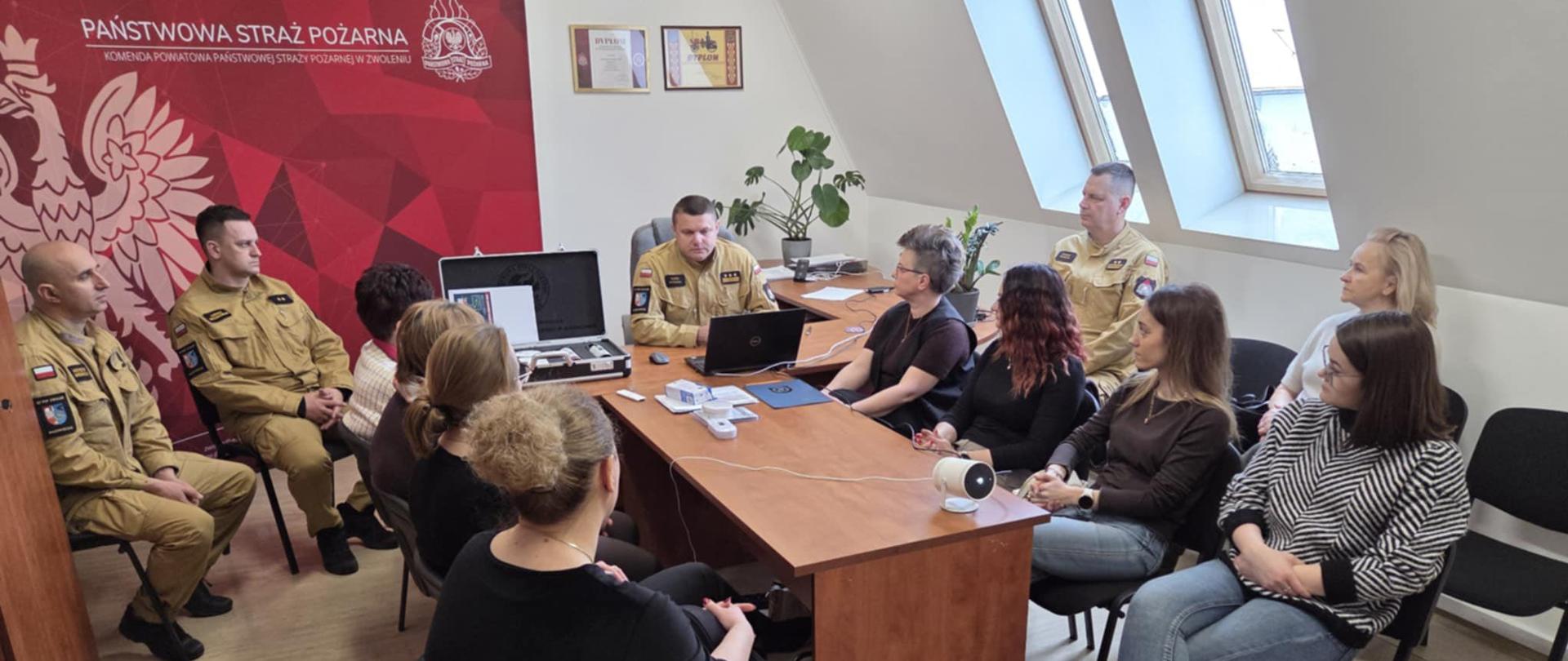 Spotkanie informacyjne w sali konferencyjnej Komendy Powiatowej Państwowej Straży Pożarnej. Przy stole siedzą strażacy w mundurach oraz grupa kobiet uczestniczących w szkoleniu. Na stole znajduje się laptop, projektor i materiały edukacyjne. W tle widoczna jest czerwona ścianka z logo Państwowej Straży Pożarnej oraz okna dachowe doświetlające pomieszczenie.