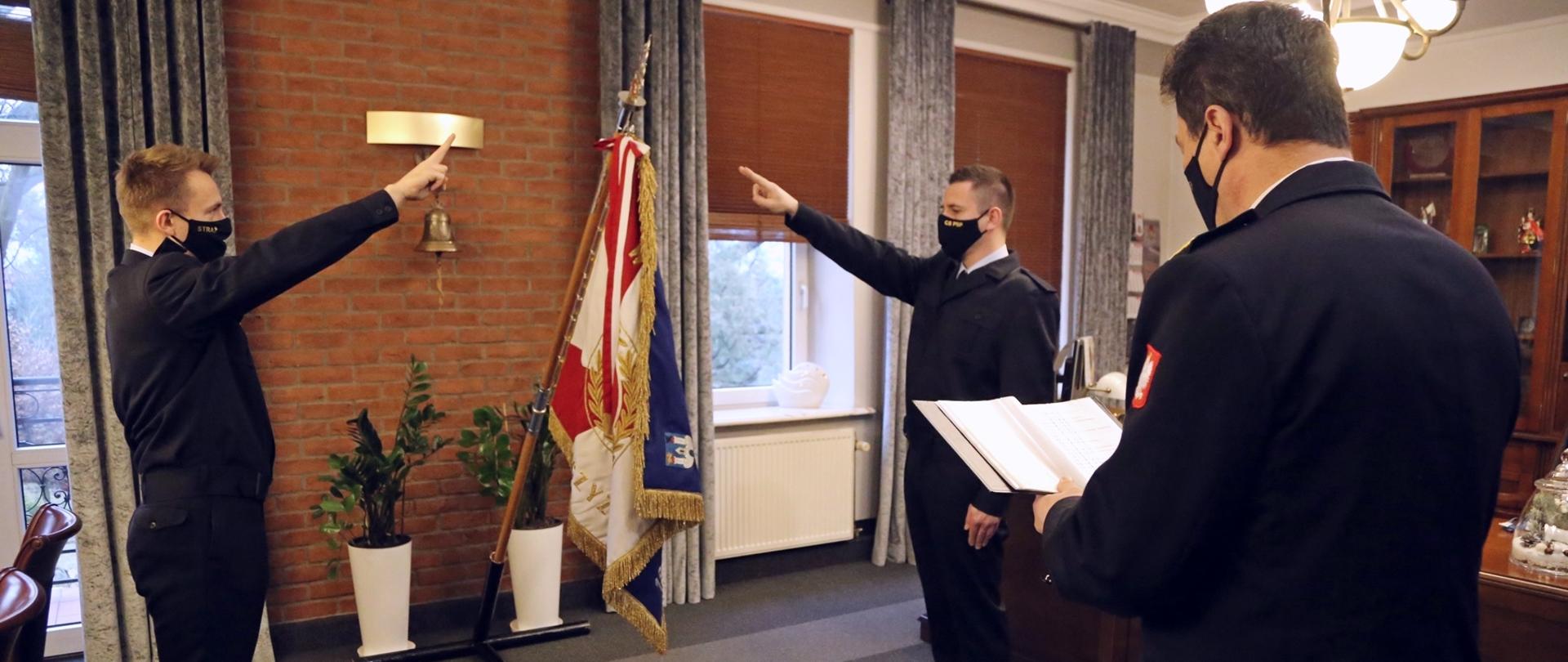 Komendant Centralnej Szkoły PSP stoi, czytając tekst roty. W głębi centralnie umieszczony stojak ze sztandarem Szkoły. Po obu stronach sztandaru stoją, z wyciągniętą lewą ręką, ślubujący strażacy