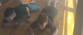 Na zdjęciu widać dzieci biorące udział w ćwiczeniach ewakuacyjnych, symulujących sytuację pożaru w zadymionym korytarzu. Poruszają się one czołgając się przy podłodze, zgodnie z zasadami bezpieczeństwa w warunkach ograniczonej widoczności i zadymienia. Na ścianach i linie zawieszone są dekoracyjne symbole płomieni, które podkreślają charakter symulacji.