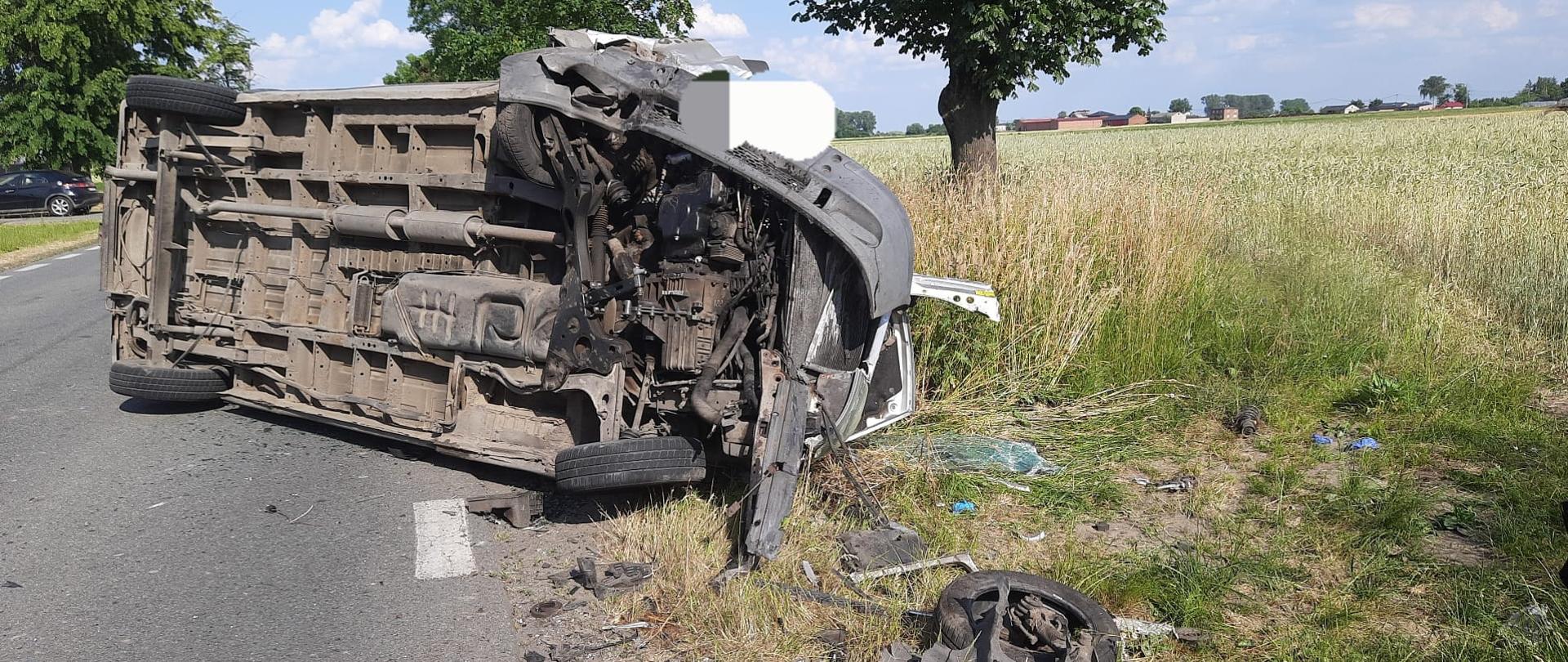 Na zdjęciu widać samochód dostawczy, który leży na boku na drodze. Widoczny jest spód pojazdu. Przed samochodem porozrzucane są elementy wyposażenia. W tle widać czarny samochód osobowy, budynki, pola i drzewa. Jest widno.