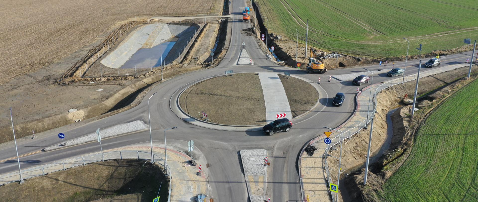 Fotografia przedstawia rondo na skrzyżowaniu dróg w otoczeniu pól uprawnych. Na zdjęciu widać kilka samochodów poruszających się po rondzie oraz maszyny budowlane pracujące przy drodze prowadzącej od ronda w głąb pola. Droga ta jest w trakcie budowy lub modernizacji, co sugerują wykopy po obu jej stronach oraz obecność sprzętu budowlanego. W tle widoczne są rozległe pola uprawne oraz linia drzew na horyzoncie. Zdjęcie jest interesujące, ponieważ pokazuje infrastrukturę drogową w trakcie budowy oraz jej integrację z otaczającym krajobrazem rolniczym.