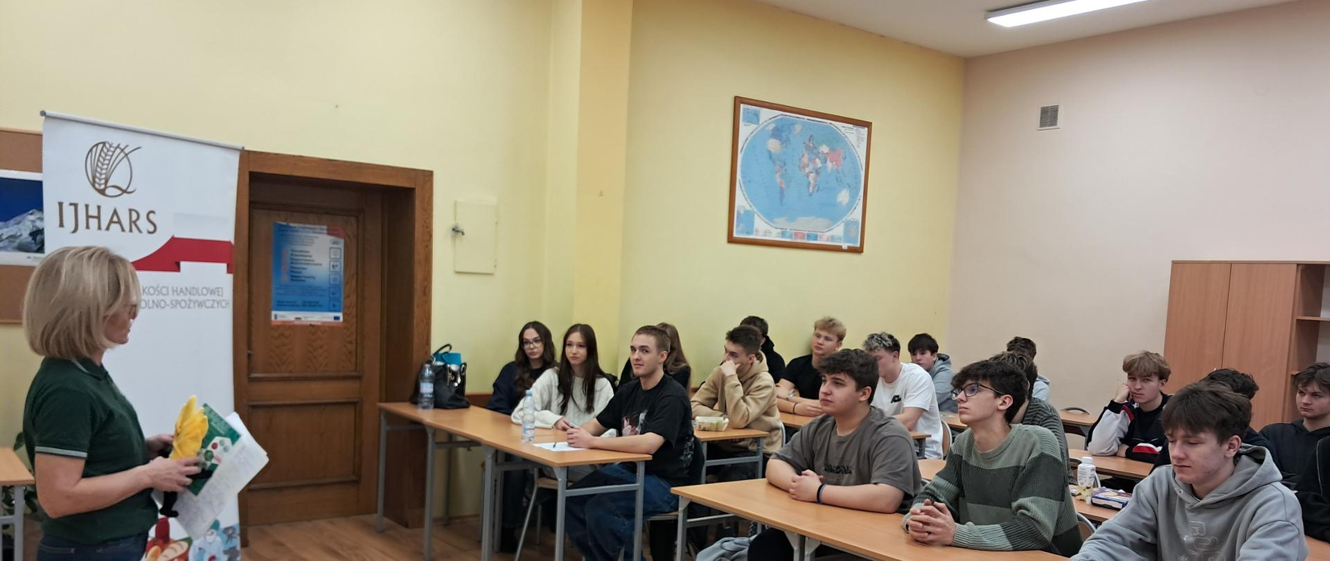 Inspektorka IJHARS w klasie z uczniami podczas lekcji, w tle logo IJHARS, uczniowie siedzą w ławkach i słuchają