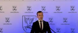 Elegancko ubrany mężczyzna przemawia na mównicy przez mikrofon na ściance Gminy Biskupiec 