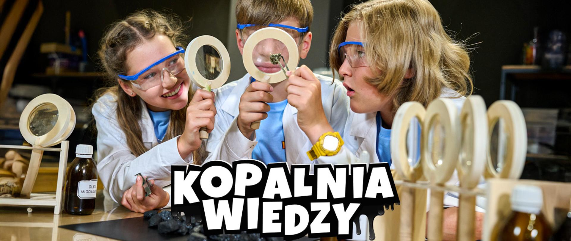 Grupa trojga dzieci w białych fartuchach i okularach ochronnych, które badają czarne kawałki węgla za pomocą lup. Po lewej stronie dziewczynka, w środku chłopiec, a po prawej drugi chłopiec. Wszyscy uśmiechają się i wyglądają na zaintrygowanych. Na pierwszym planie widać duży, biały napis „KOPALNIA WIEDZY”.