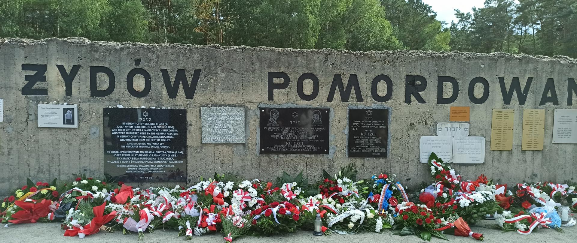 Zajecie fragmentu muru Muzeum byłego niemieckiego Obozu Zagłady Kulmhof w Chełmnie nad Nerem z napisem "PAMIĘCI ŻYDÓW ZAMORDOWANYCH W CHEŁMNIE 1941-1945". Pod murem duża ilość kwiatów złożonych podczas uroczystości.