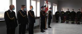 na zdjęciu w pomieszczeniu sali konferencyjnej komendy stoi trzyosobowy poczet flagowy, strażacy ubrani są w mundury galowe ze sznurem, na głowie maja czapkę rogatywkę i czarne rękawiczki. Z lewej strony pocztu stoi komendant powiatowy PSP w Łowiczu mł. bryg. Arkadiusz Makowski, zastępca komendanta powiatowego mł. bryg. Rafał Garbarz, p.o. dowódcy JRG kpt. Jakub Wojda, strażacy ubrani są w mundury galowe ze sznurem. Z prawej strony pocztu stoją funkcjonariusze pełniący służbę w JRG PSP w Łowiczu, strażacy ubrani są w ubrania koszarowe. Wszyscy mają maseczki chirurgiczne. Zdjęcie w formacie panoramicznym. 
