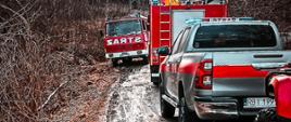 Zdjęcie przedstawia samochody strażackie na drodze szutrowej. Jeden z pojazdów należy do jednostki OSP a przy nim ustawione w przeciwnym kierunku są: samochód gaśniczy Man oraz Toyota Hilux z podpiętą przyczepką na której znajduje się quad. Te pojazdy należą do JRG Ustrzyki Dolne.
