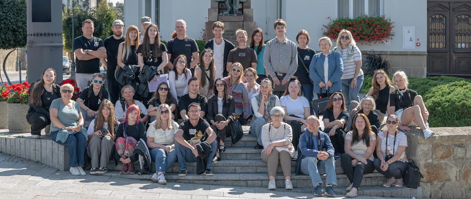 Zdjęcie przedstawia grupę słuchaczy i nauczycieli Studium pozujących na tle budynku oraz figurze stojącej na cokole. 