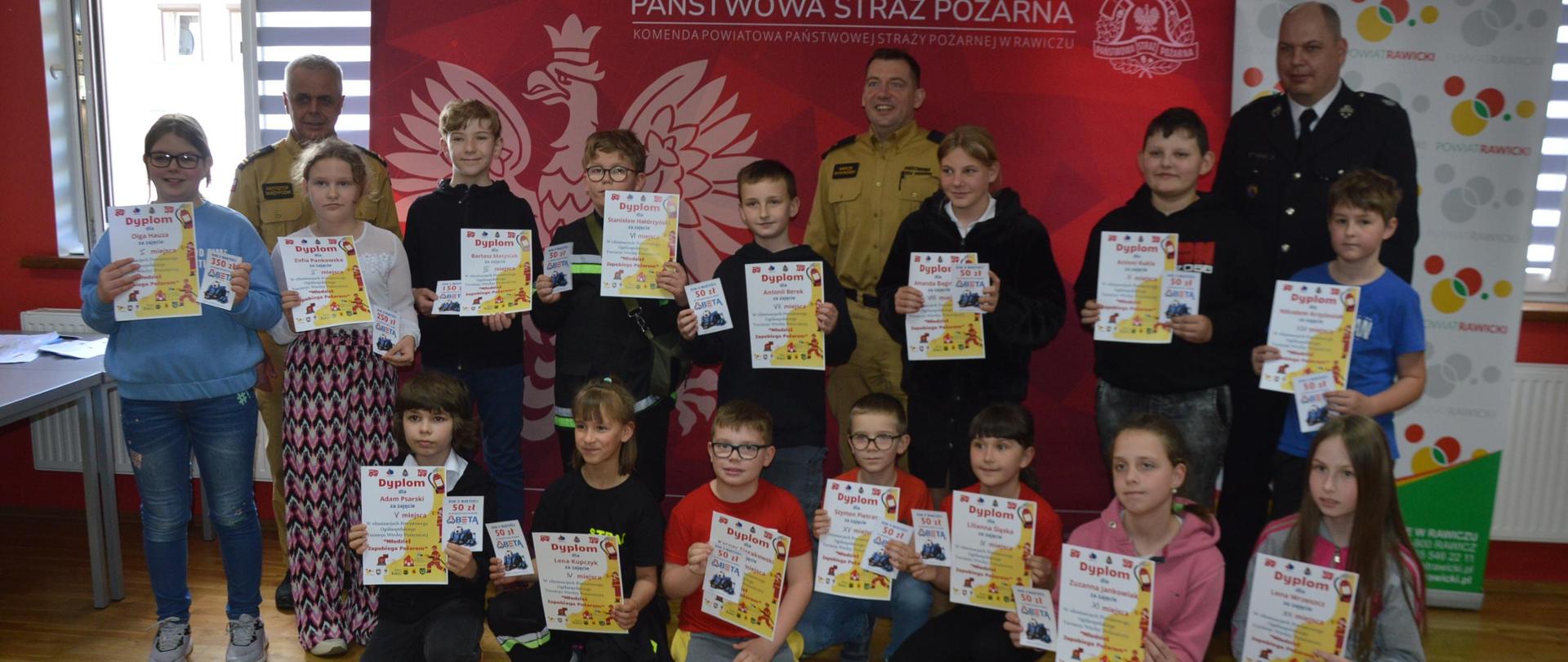 Świetlica komendy. Na tle ścianki promocyjnej wraz z organizatorami do pamiątkowego zdjęcia pozują uczestnicy eliminacji z najmłodszej grupy wiekowej. Trzymaja w rękach dyplomy i upominki. 