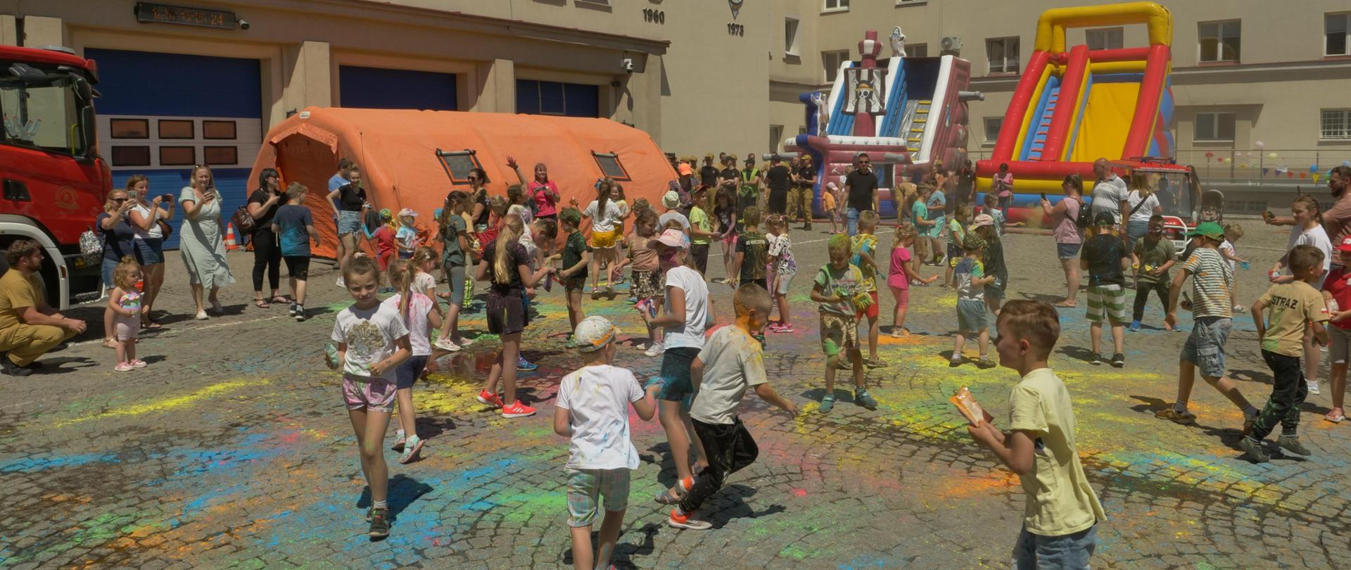 Plac wewnętrzny Szkoły Aspirantów PSP w Krakowie. Na środku dzieci w czasie festiwalu kolorów.. W tle budynki Szkoły, dmuchane ślizgawki, balony i pomarańczowy namiot pneumatyczny. 