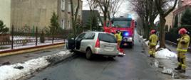Na zdjęciu widać samochód osobowy, który uderzył w przydrożne drzewo. Za rozbitym pojazdem widać jak strażacy udzielają pomocy poszkodowanemu, na jezdni przy strażakach leży torba medyczna. Za nimi, na drodze stoi samochód pożarniczy