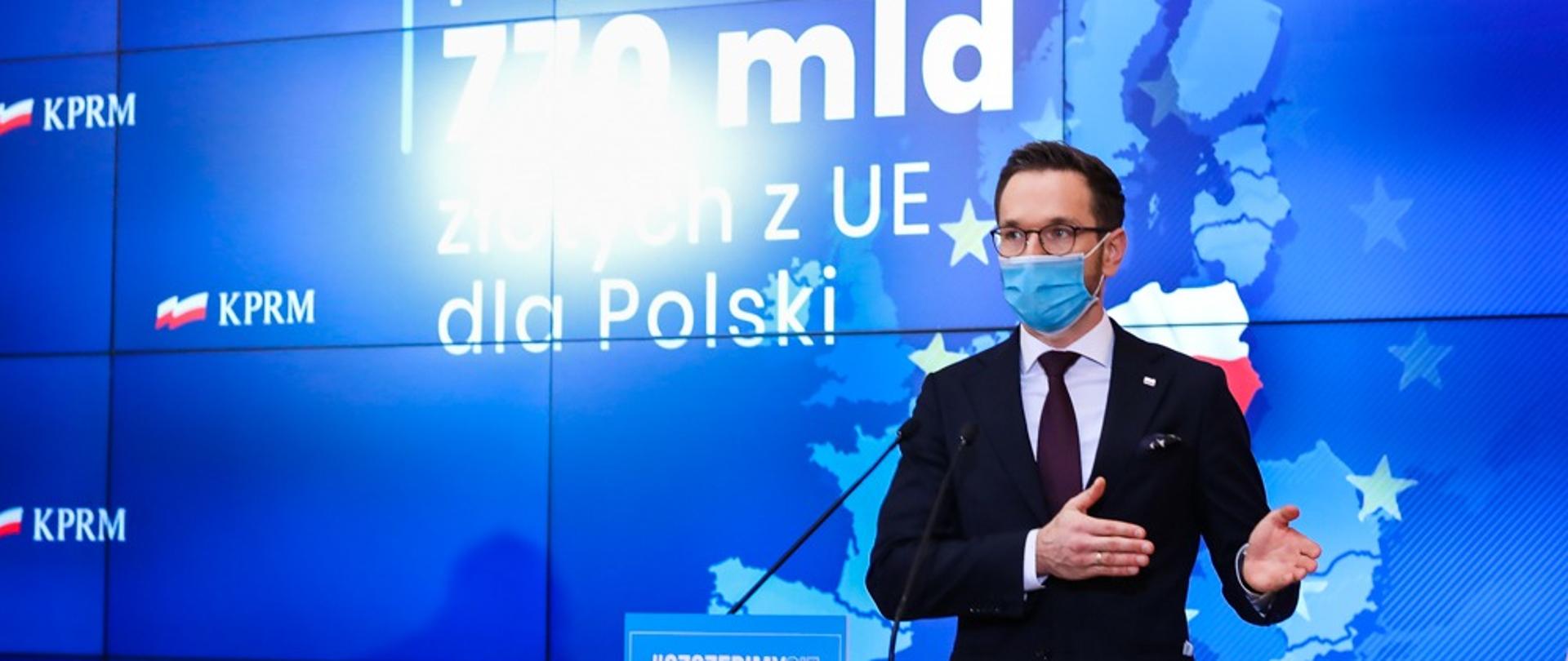 Wiceminister Waldemar Buda stoi w maseczce przed mównicą. Za plecami ekran z napisem: "Ponad 770 mld złotych z UE dla Polski".