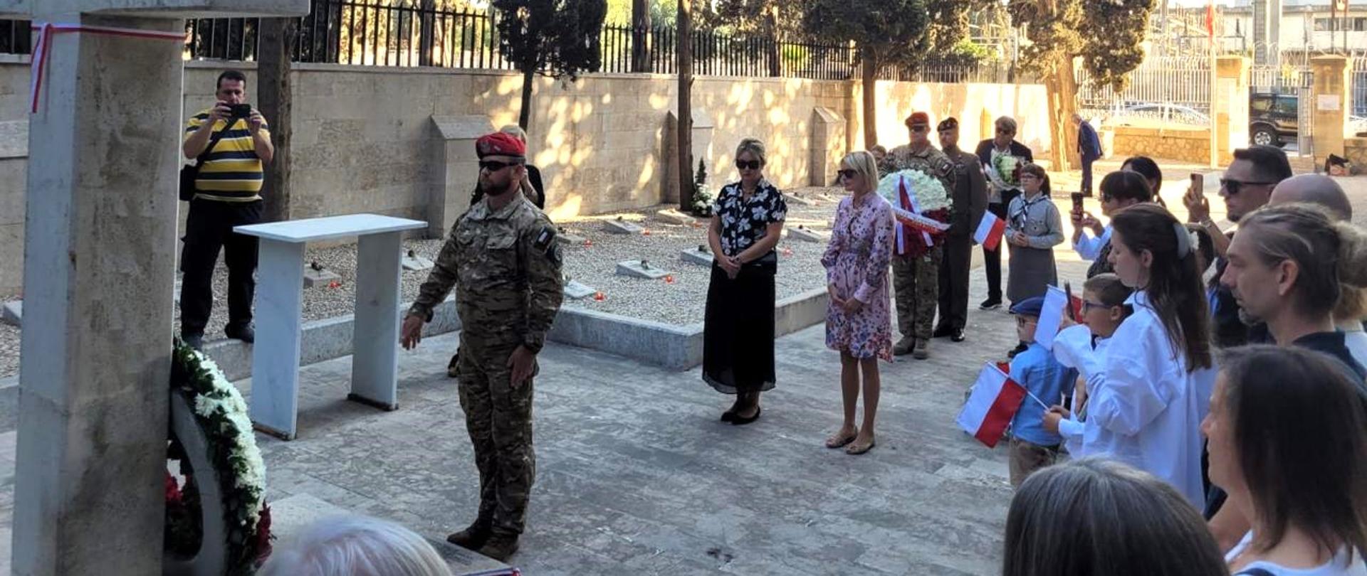 Polski Cmentarz Wojenny w Bejrucie. Po lewej betonowy krzyż, który znajduje się na środku. Żołnierz w trakcie składania wieńca, W środku Ambasador RP w Bejrucie, Aleksandra Bukowska-McCabe i Konsul RP, Aneta Ananicz. Na prawo tłum przybyłych ob. RP i członkowie rodzin, pochowanych na Cmentarzu..