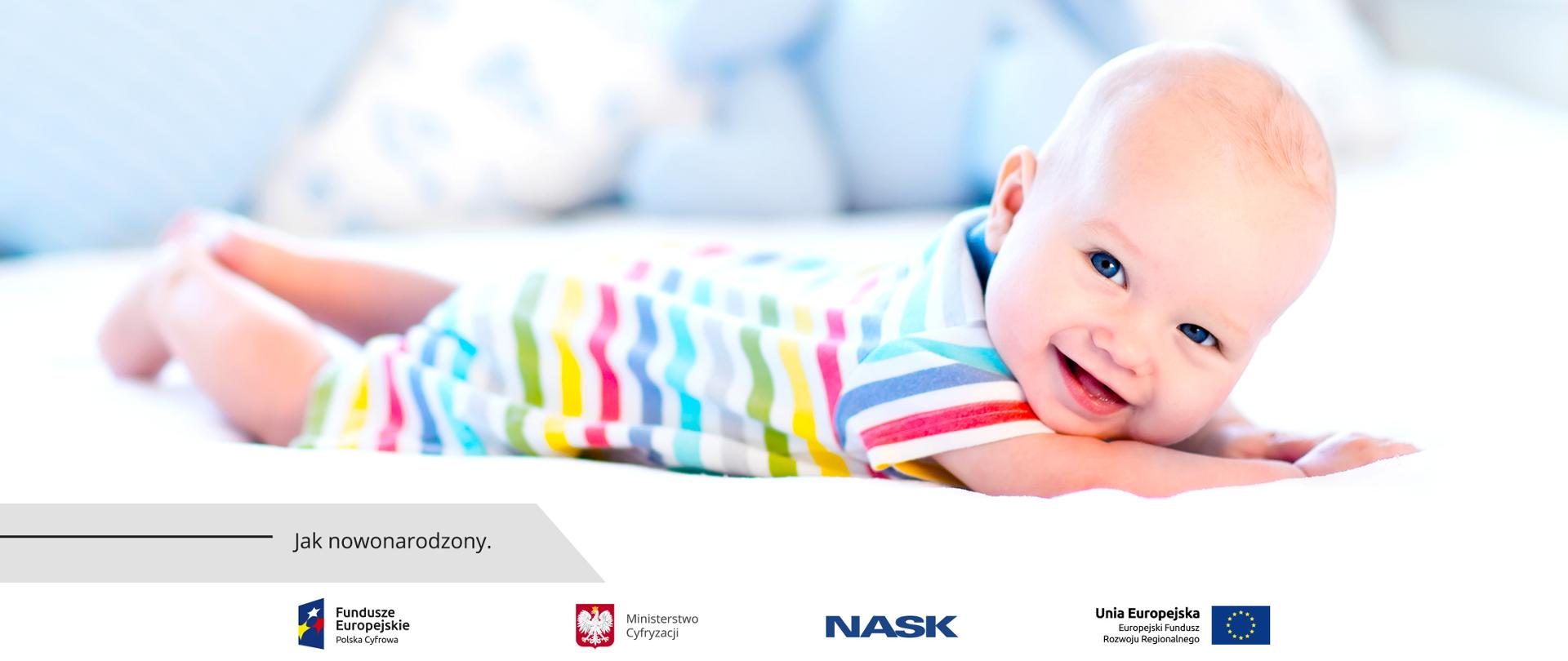 Uśmiechnięty niemowlak leżący na łóżku, na brzuszku, ubrany w kolorowe śpioszki.