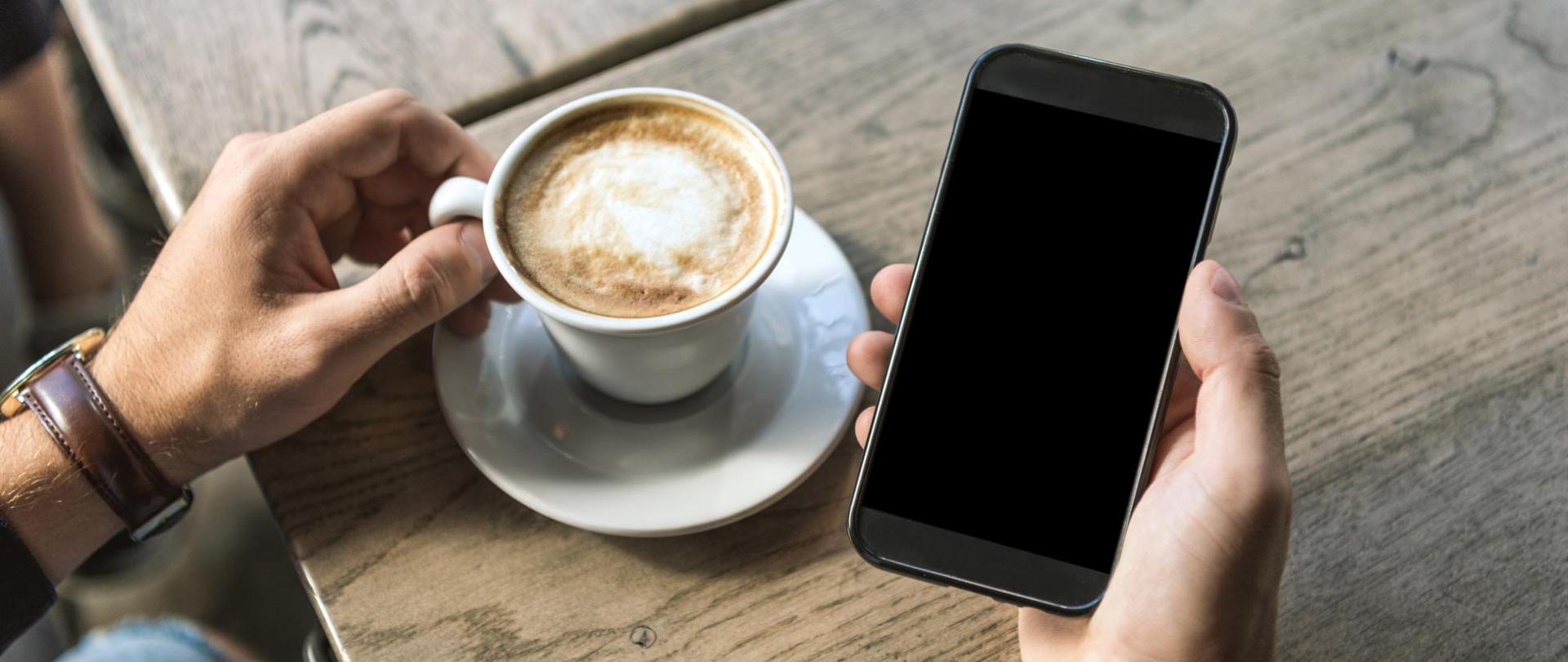 Na zdjęciu widać dłonie mężczyzny. W jednej trzyma smartfon a w drugiej filiżankę kawy.