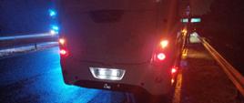 Na poboczu drogi przy barierkach stoi autobus, widać jego tył. W tle widać niebieskie światła alarmowe. Wokół jest ciemno.