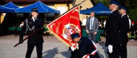 Zdjęcie zrobione na zewnątrz, podczas uroczystego apelu. Strażak trzyma pochylony sztandar jednostki OSP. Materiał sztandaru całuje klęczący strażak ochotnik. 