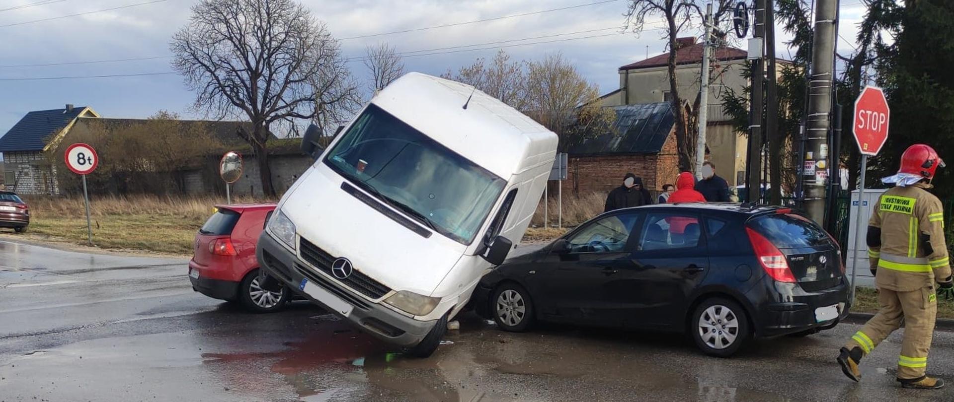 Na zdjęciu przedstawiono trzy samochody biorące udział w kolizji. Auta osobowe: czerwony Volkswagen i czarny Hyundai stoją na jezdni pod katem 90º w stosunku do siebie. Miedzy nimi, opierając się tyłem na masce Volkswagena stoi dostawczy mercedes Sprinter. Po prawej stronie stoi ratownik a za samochodami grupa świadków zdarzenia.