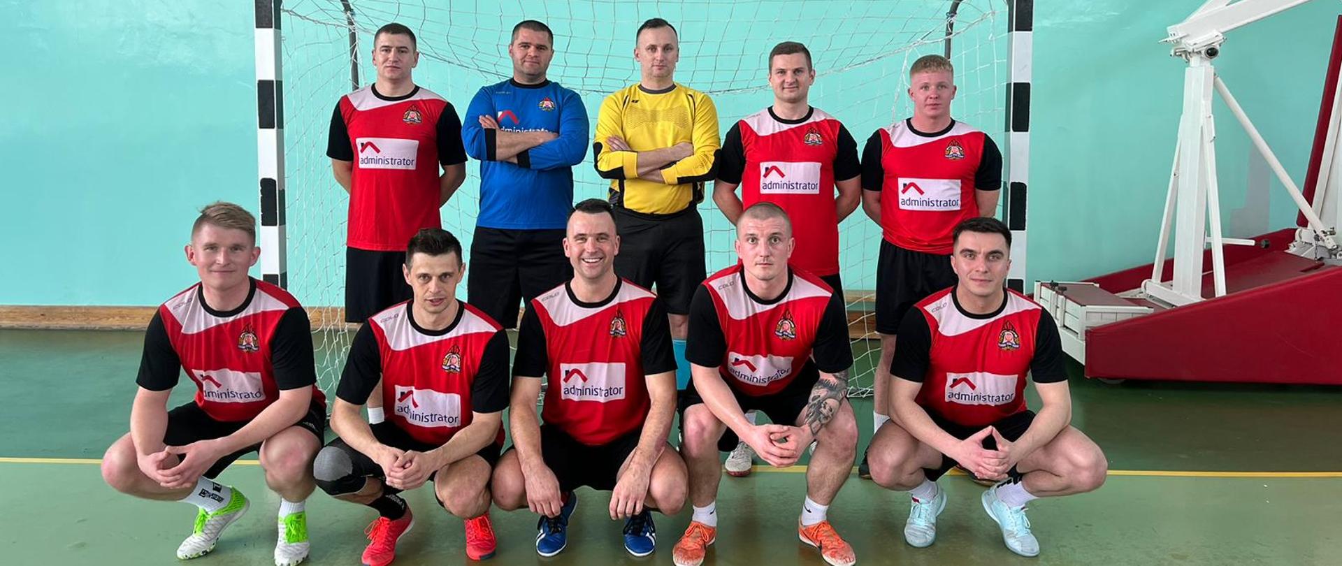 Zdjęcie wykonane na terenie obiektu sali sportowej w Sochaczewie podczas rozgrywek strefowych strażaków w piłce nożnej. Na boisku widać ustawioną do pamiątkowego zdjęcia zwycięską drużynę z komendy miejskiej Państwowej Straży Pożarnej w Radomiu. Zawodnicy ubrani są w stroje sportowe w kolorze czerwono - czarno – białym z wyjątkiem dwóch bramkarzy którzy posiadają stroje w kolorach niebiesko – czarnym oraz żółto – czarnym. 