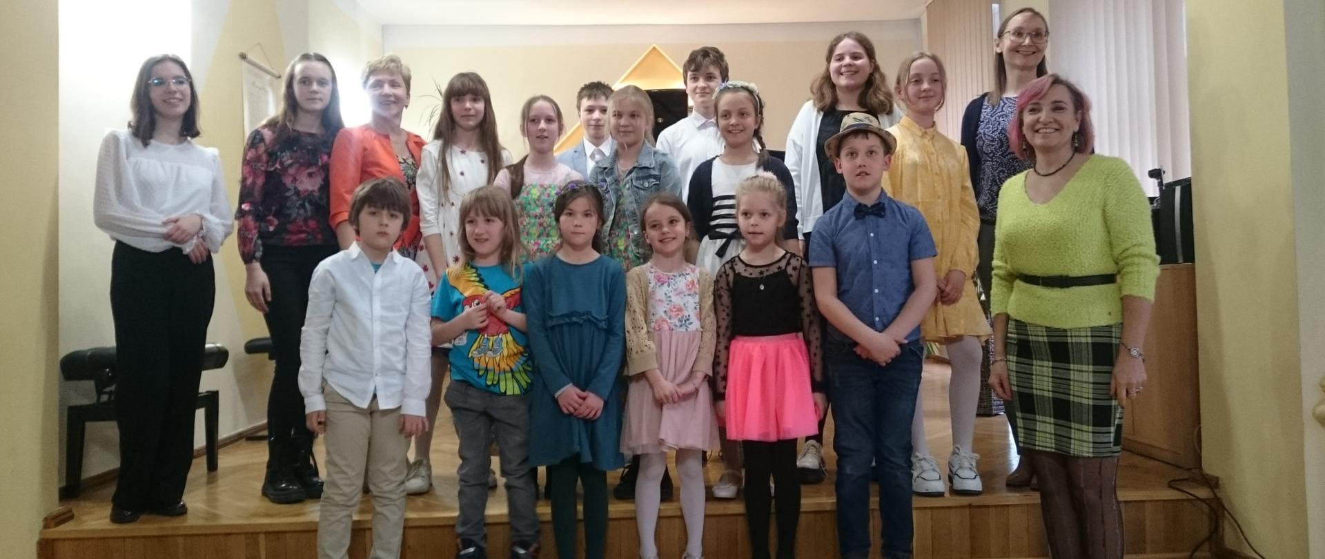 Zdjęcie grupowe uśmiechniętych uczniów wraz z nauczycielami P. J. Kocyan, A. Strzygocką biorących udział w koncercie "Mój ulubiony utwór" w dniu 19.04.2023, Zdjęcie wykonane na scenie auli PSM I st. w Kłodzku