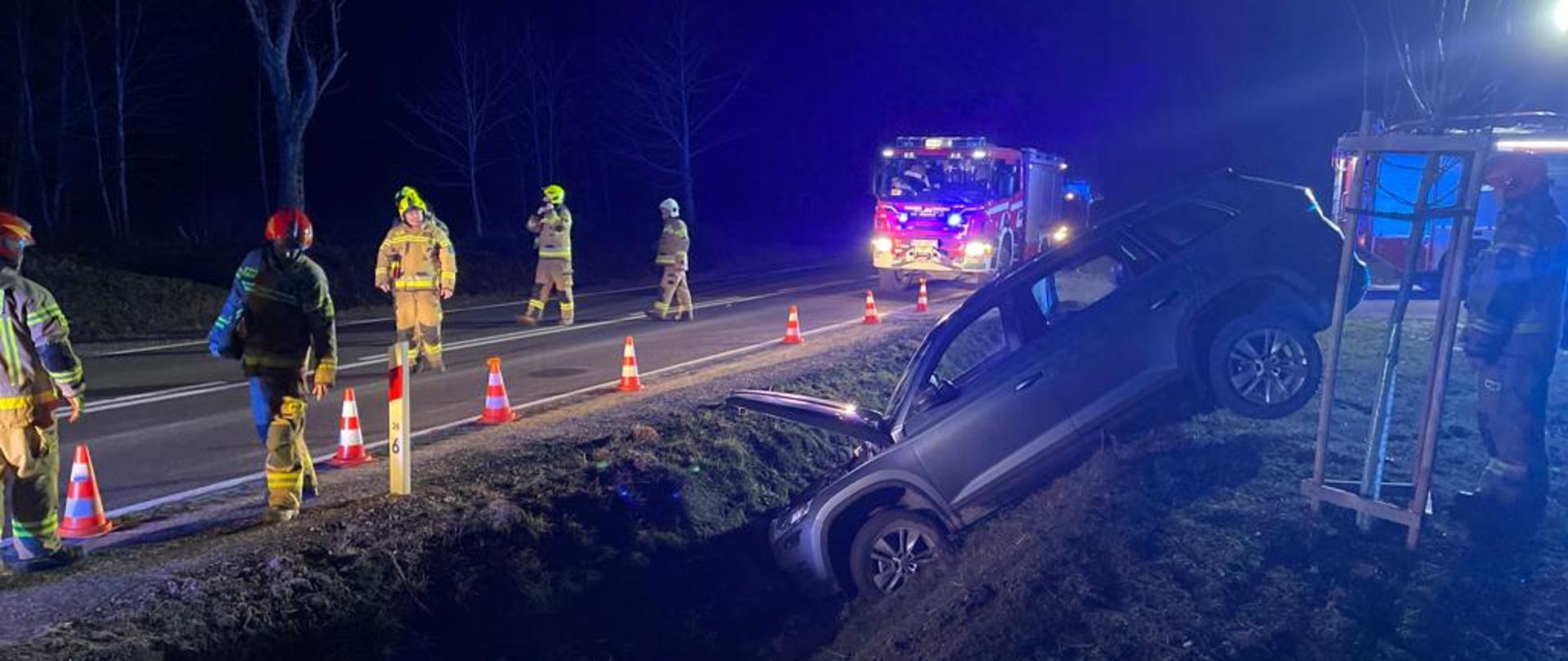 Zdjęcie zrobione nocą, Strażacy na drodze dalej drogę oświetla samochód strażacki. Z prawej strony samochód przodem w rowie.