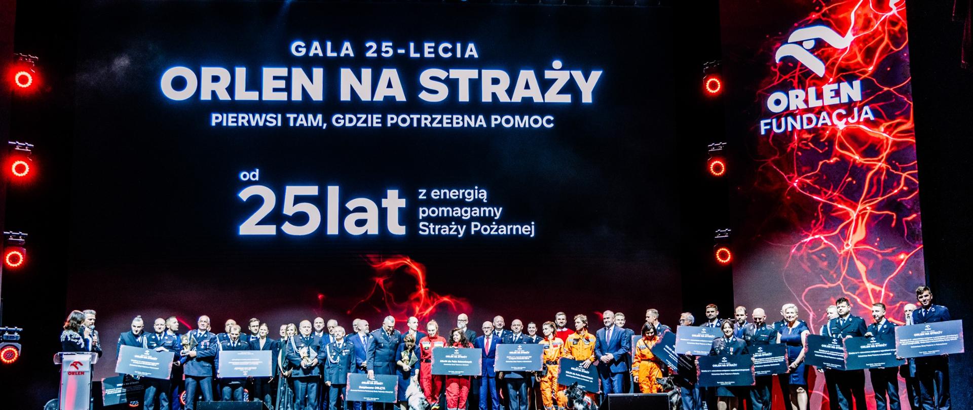 Szerokie ujęcie przedstawia wielką scenę Teatru Wielkiego – Opery Narodowej w Warszawie z ustawioną na niej liczną grupą kilkudziesięciu laureatów oraz reprezentantów służb. W centralnej części sceny stoją obok siebie strażacy Państwowej Straży Pożarnej w wyjściowych, ciemnogranatowych mundurach galowych z pagonami, ratownicy w jasnopomarańczowych i czerwono-żółtych kombinezonach specjalistycznych oraz przedstawiciele organizacji w eleganckich strojach cywilnych, takich jak garnitury i ciemne sukienki. Wielu wyróżnionych trzyma przed sobą duże, prostokątne pamiątkowe czeki z logo organizatora. W tle wznosi się imponujący, gigantyczny ekran LED o czarnym tle z rozświetlonymi na czerwono grafikami imitującymi wyładowania energii. Na ekranie widnieją duże, białe napisy: „GALA 25-LECIA ORLEN NA STRAŻY”, poniżej podtytuł „PIERWSI TAM, GDZIE POTRZEBNA POMOC”, a pod nim napis „25 lat z energią pomagamy Straży Pożarnej”. Po prawej stronie ekranu umieszczono pionowy napis „ORLEN FUNDACJA” wraz z logo. Scena spowita jest w profesjonalnym oświetleniu teatralnym z jasnymi snopami białych reflektorów z góry oraz ciepłą, czerwoną łuną u dołu. Ze zdjęcia bije podniosła atmosfera, duma oraz poczucie wspólnoty i uroczystego zwieńczenia lat owocnej współpracy.