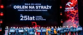 Szerokie ujęcie przedstawia wielką scenę Teatru Wielkiego – Opery Narodowej w Warszawie z ustawioną na niej liczną grupą kilkudziesięciu laureatów oraz reprezentantów służb. W centralnej części sceny stoją obok siebie strażacy Państwowej Straży Pożarnej w wyjściowych, ciemnogranatowych mundurach galowych z pagonami, ratownicy w jasnopomarańczowych i czerwono-żółtych kombinezonach specjalistycznych oraz przedstawiciele organizacji w eleganckich strojach cywilnych, takich jak garnitury i ciemne sukienki. Wielu wyróżnionych trzyma przed sobą duże, prostokątne pamiątkowe czeki z logo organizatora. W tle wznosi się imponujący, gigantyczny ekran LED o czarnym tle z rozświetlonymi na czerwono grafikami imitującymi wyładowania energii. Na ekranie widnieją duże, białe napisy: „GALA 25-LECIA ORLEN NA STRAŻY”, poniżej podtytuł „PIERWSI TAM, GDZIE POTRZEBNA POMOC”, a pod nim napis „25 lat z energią pomagamy Straży Pożarnej”. Po prawej stronie ekranu umieszczono pionowy napis „ORLEN FUNDACJA” wraz z logo. Scena spowita jest w profesjonalnym oświetleniu teatralnym z jasnymi snopami białych reflektorów z góry oraz ciepłą, czerwoną łuną u dołu. Ze zdjęcia bije podniosła atmosfera, duma oraz poczucie wspólnoty i uroczystego zwieńczenia lat owocnej współpracy.