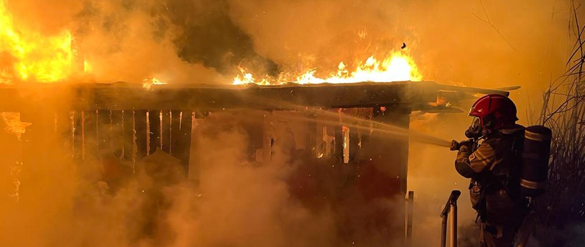 Zdjęcie przedstawia strażaka gaszącego pożar obiektu drewnianego