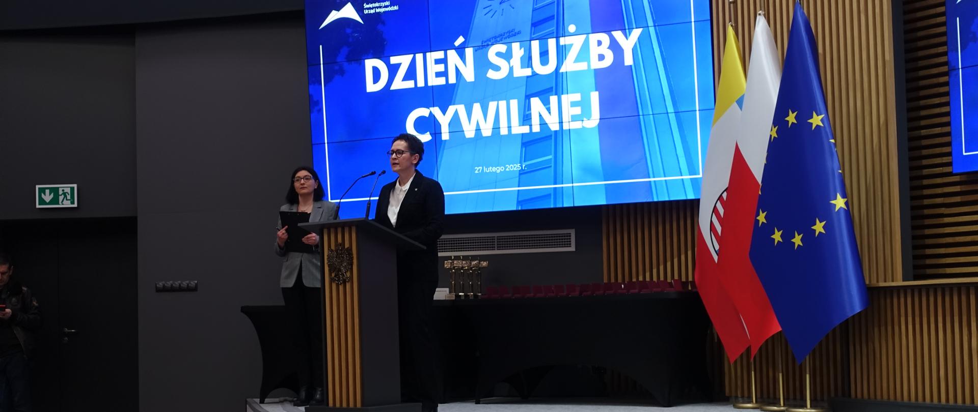 Zdjęcie przedstawia uroczyście przygotowaną scenę Sali konferencyjnej w Świętokrzyskim Urzędzie Wojewódzkim. Przy konsoli stoi kobieta i wygłasza przemówienie, obok zaś stoi druga kobieta trzymająca notatnik. Za kobietami widoczny jest ekran z napisem Dzień Służby Cywilnej.