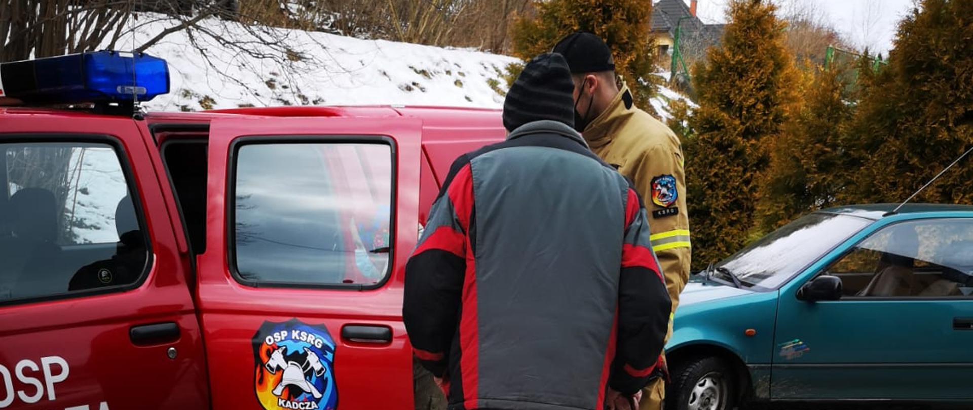 Osoba korzystająca z transportu o lasce zbliża się do samochodu operacyjnego OSP Kadcza. Obok stoi strażak.