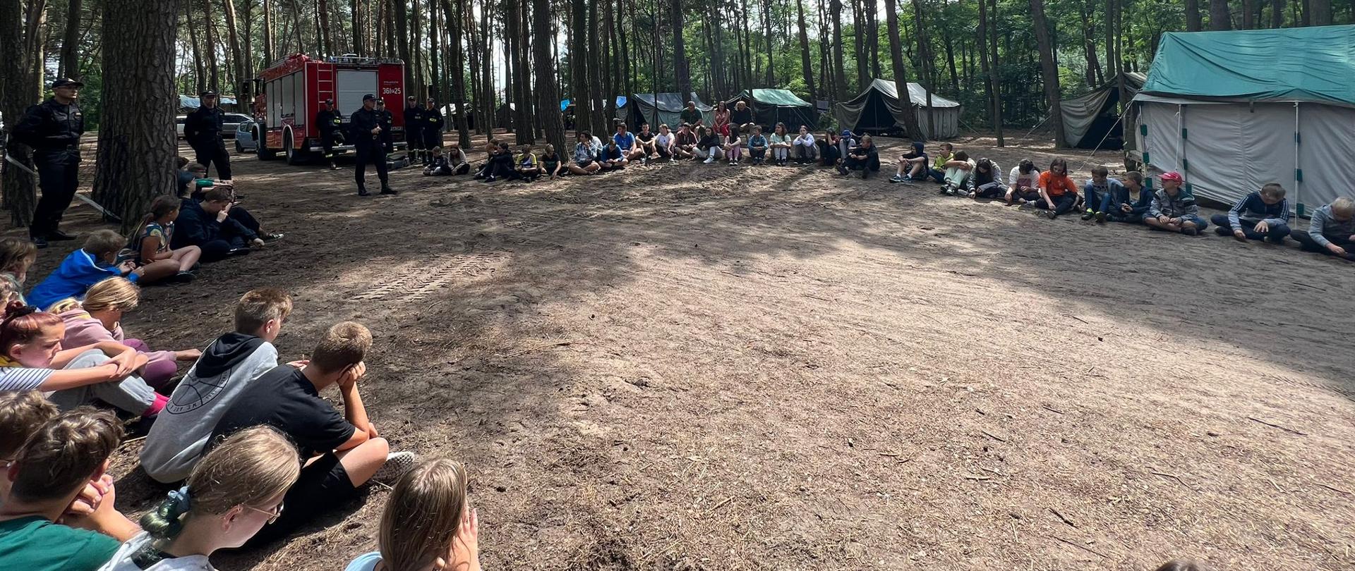 Zdjęcie przedstawia obozowiczów, którzy siedząc na ziemi tworzą duże koło i słuchają prelekcji strażaków. W tle drzewa, namioty oraz wóz strażacki.