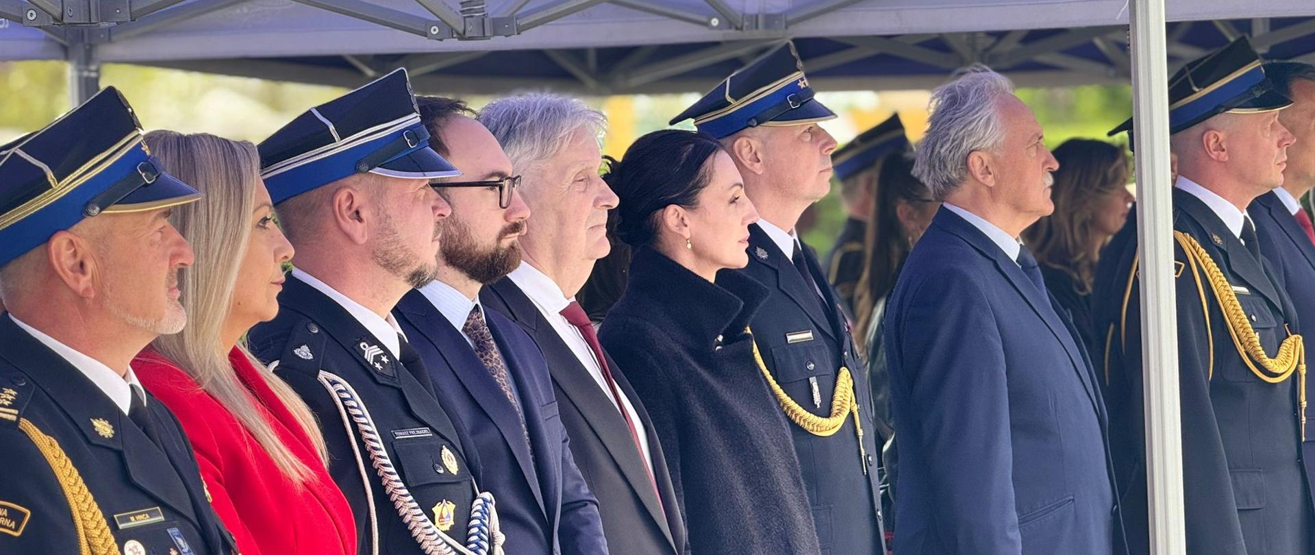 Zdjęcie przedstawia grupę osób stojących w rzędzie podczas oficjalnej uroczystości. Postacie ustawione są profilem, patrząc w jednym kierunku.