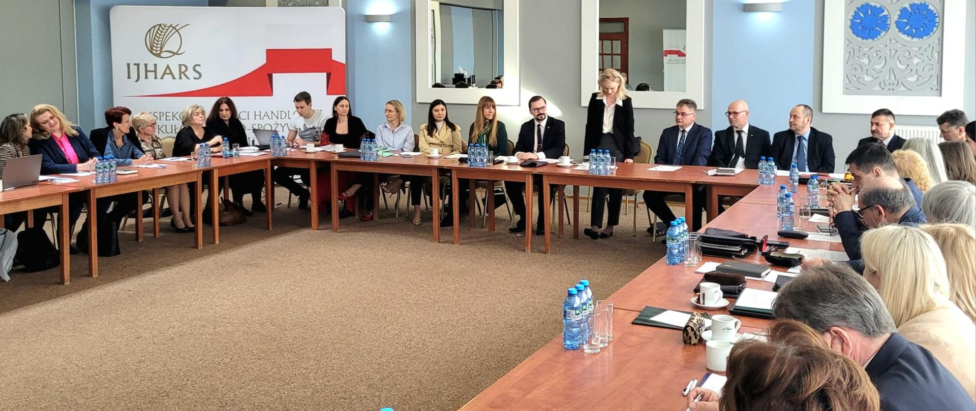 Ogólnopolska narada IJHARS, wnętrze sali konferencyjnej, stoły za którymi siedzą uczestnicy spotkania ustawione w podkowę, w tle ścianka reklamowa z logo IJHARS i flagą biało- czerwoną.