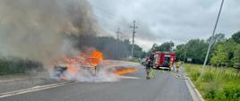 Na zdjęciu widać palący się samochód osobowy. Strażak zabezpieczony w aparat ochrony dróg oddechowych gasi ogień.