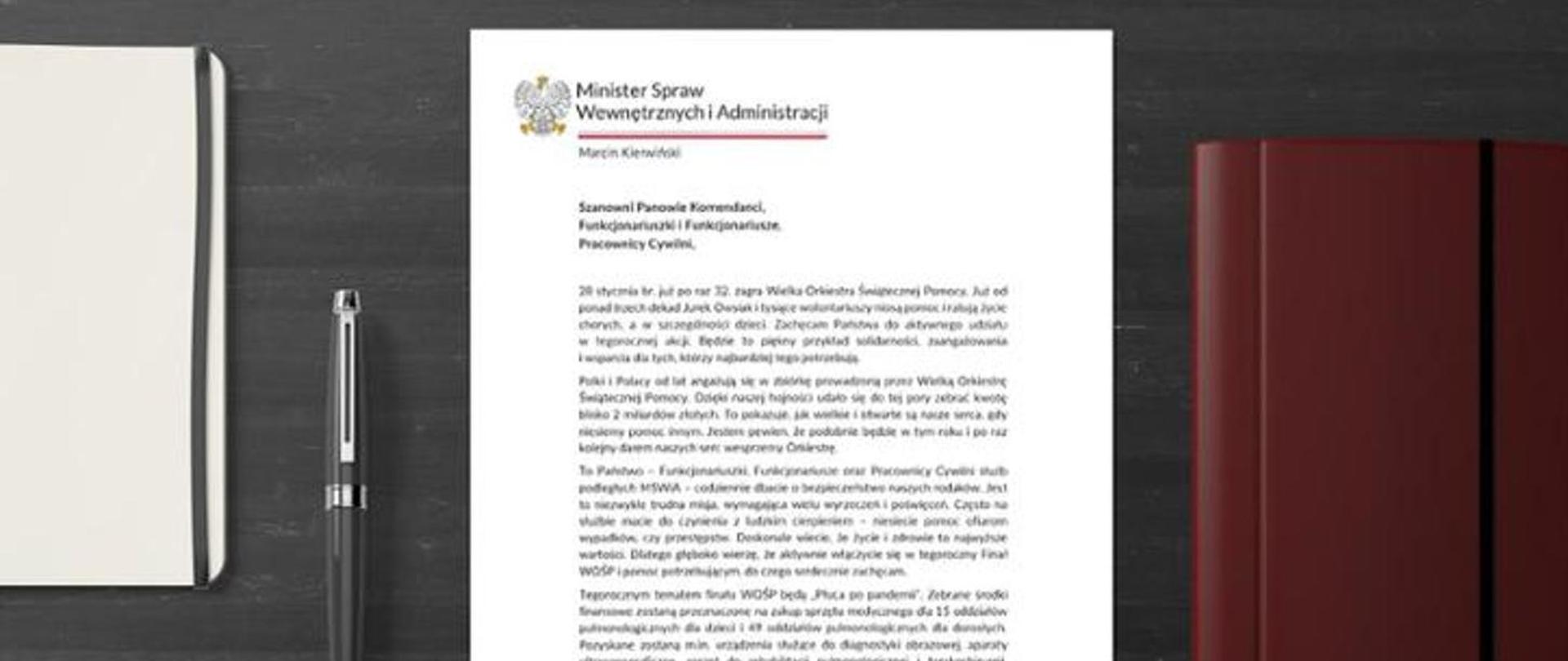 Szary blat, na którym leży list spisany na białej kartce, długopis oraz dwa notatniki.