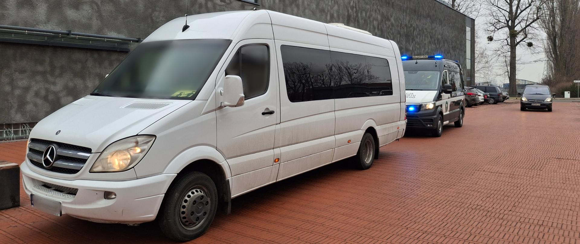 Biały autobus pasażerski marki Mercedes-Benz Sprinter zaparkowany na placu wyłożonym czerwoną kostką brukową. Zaraz za nim stoi furgon Inspekcji Transportu Drogowego (ITD) z włączonymi niebieskimi sygnałami świetlnymi. W tle widoczny jest nowoczesny budynek o szarej, chropowatej elewacji z podłużnymi oknami oraz wysokie drzewa bez liści.
