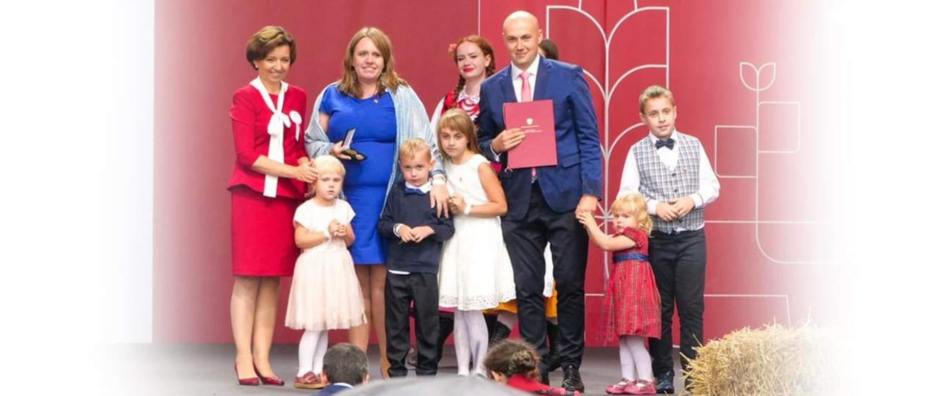Zdjęcie grupowe, podobne do zdjęcia rodzinnego. Z lewej strony ubrana w czerwony kostium, minister Maląg. Obok niej nagrodzona rodzina. Tata, mama oraz pięcioro dzieci.
