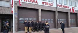 Ujęcia z chwili podnoszenia flagi lub składania honorów. Oficerowie przy maszcie (z lewej) oraz oficer na prawej flance salutują (podnoszą prawą dłoń do nakrycia głowy). Strażacy w szeregu również salutują. Napis na fasadzie jest w pełni widoczny: "PAŃSTWOWA STRAŻ POŻARNA".