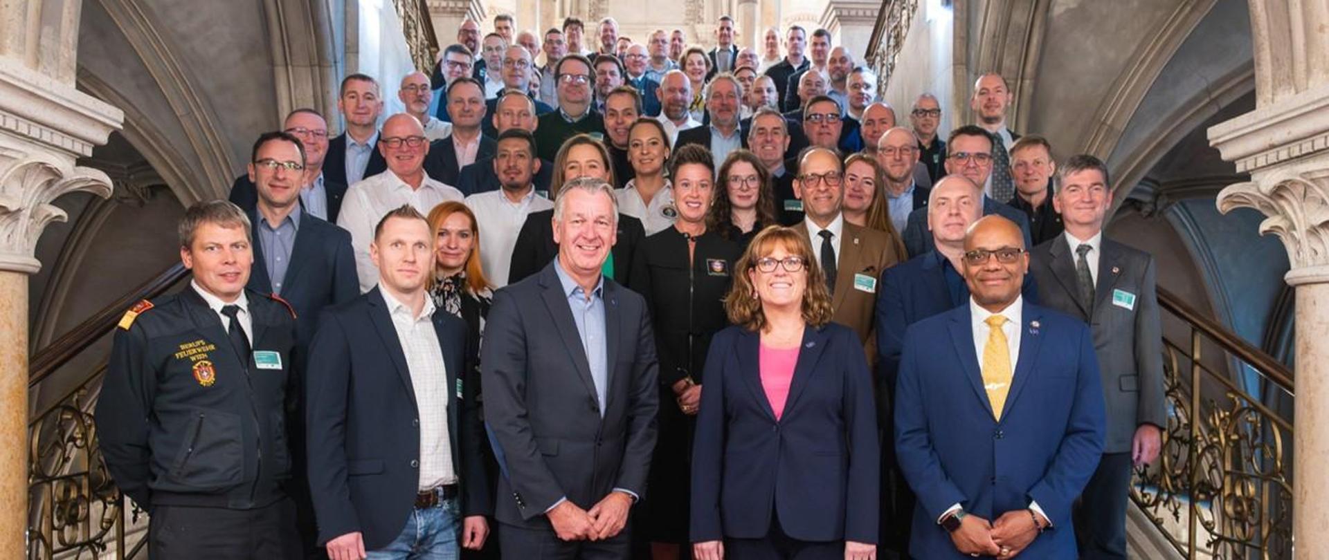 Duża grupa uczestników konferencji stoi na schodach w zabytkowym wnętrzu. W pierwszym rzędzie znajdują się osoby w strojach służbowych i garniturach. W tle widać ozdobne kamienne kolumny i balustrady.