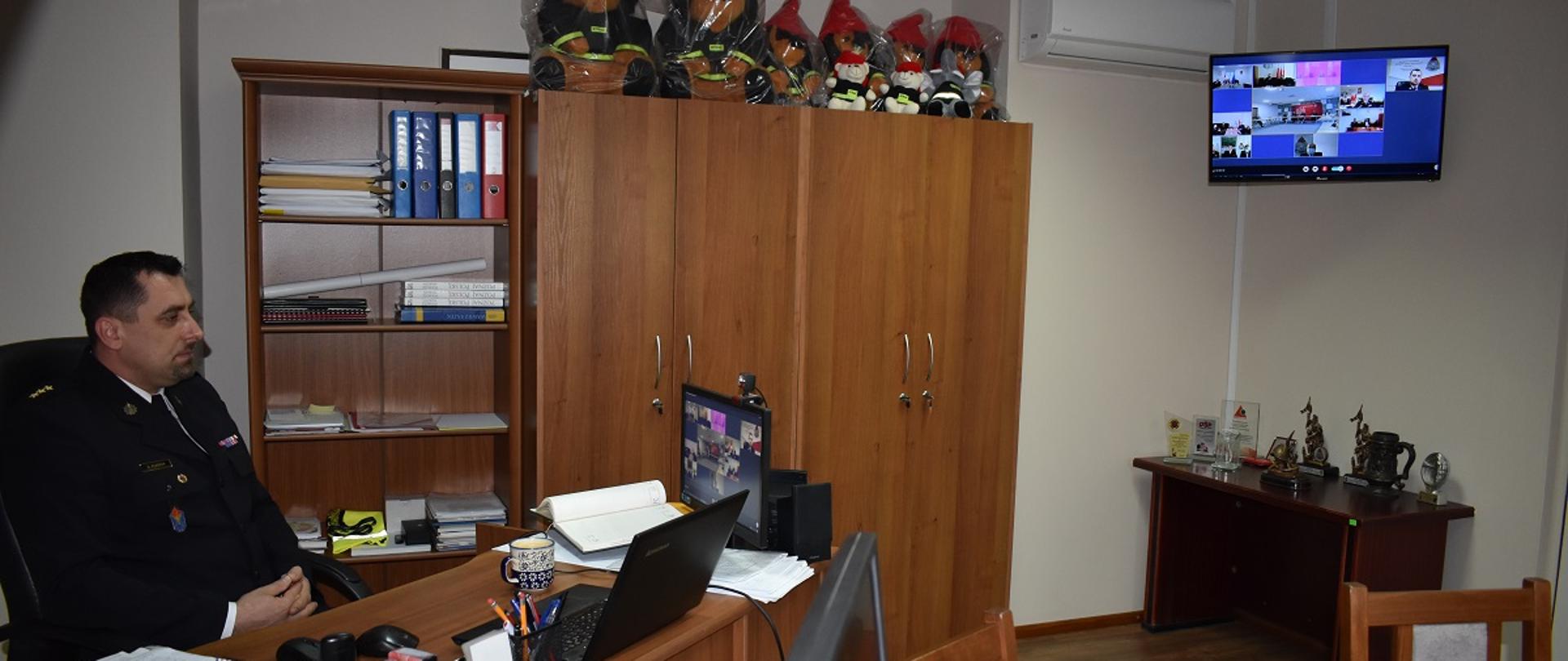 Zastępca Komendanta Powiatowego PSP siedzi za biurkiem. Na monitorze komputerowym znajdującym się na biurku oraz, w tle, telewizorze powieszonym na ścianie widać innych uczestników odprawy która odbywa się w formie wideokonferencji