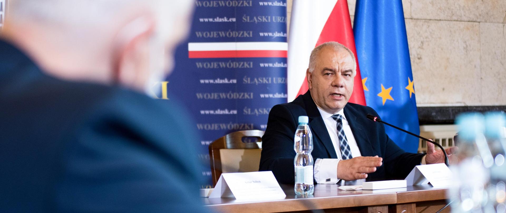 Wicepremier Jacek Sasin podczas spotkania w Katowicach. Wicepremier siedzi za stołem prezydialnym. Przed nim Dominik Kolorz. Wicepremier przemawia.