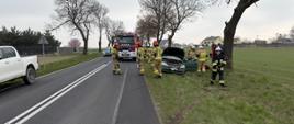 Uszkodzony samochód poza drogą wokół strażacy oraz samochody ratownicze