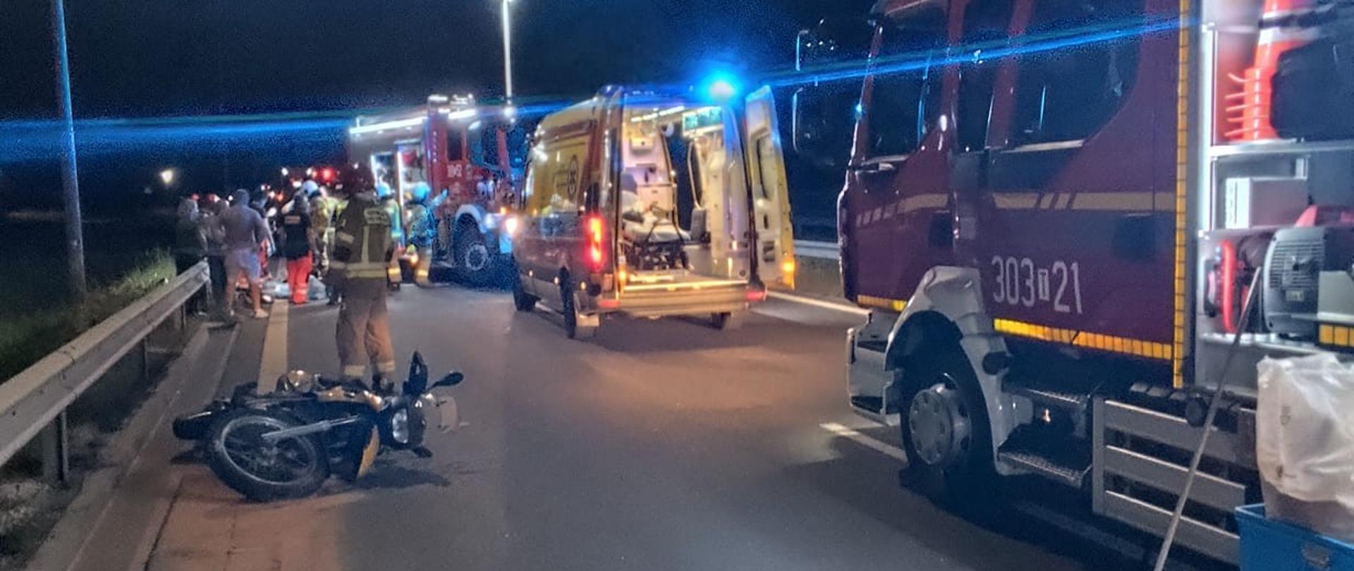 Zdjęcie przedstawia miejsce wypadku: z lewej strony, na jezdni leży zniszczony motocykl, a zanim grupa ratowników. Po prawej stronie zaś zaparkowały samochody ratownicze: kolejno samochód pożarniczy, ambulans Państwowego Ratownictwa Medycznego i kolejny samochód pożarniczy.