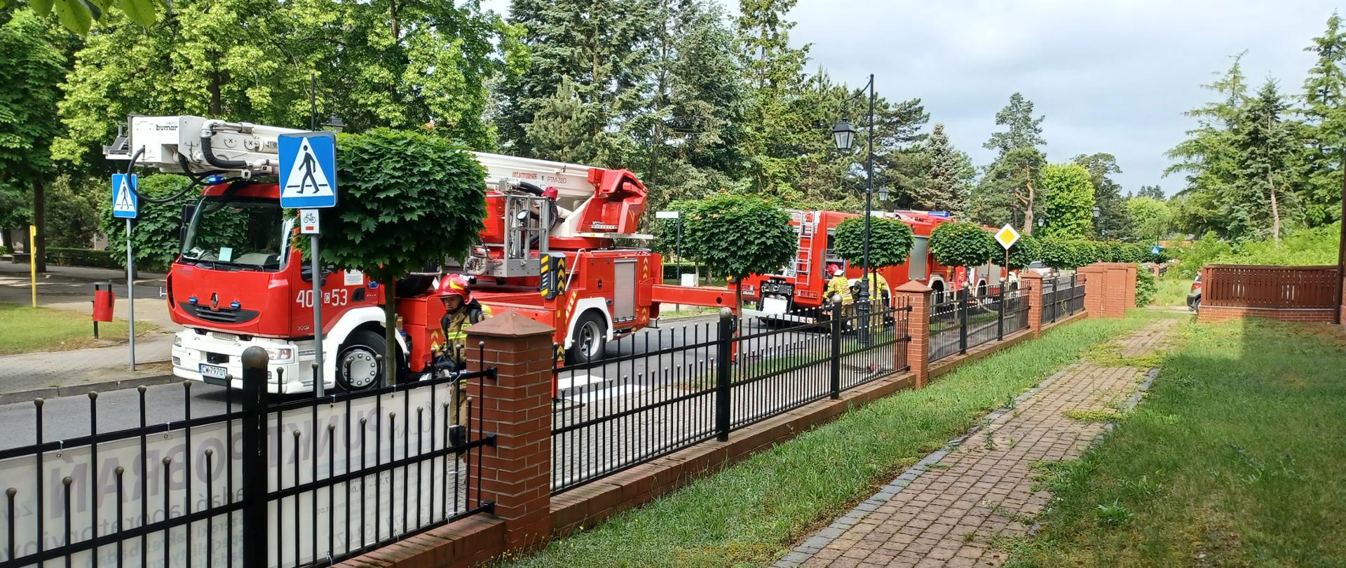 Na zdjęciu widać stojące na asfaltowej drodze koloru szarego trzy czerwone pojazdy pożarnicze w tym jeden podnośnik z drabiną. Wzdłuż drogi miejskiej usytuowane są drzewa koloru zielonego oraz oznakowanie drogowe, przejście dla pieszych i droga z pierwszeństwem przejazdu. Przy samochodach stoją strażacy ubrani w piaskowe umundurowanie i czerwone hełmy, którzy wyjmują sprzęt ze skrytek samochodowych. Zdjęcie zrobiono z ternu posesji, na której odbywają się ćwiczenia. Posesja jest ogrodzona ozdobnym ogrodzeniem wykonanym z cegły oraz czarnymi dekoracyjnymi metalowymi przęsłami. Pogoda pochmurna bez opadów.