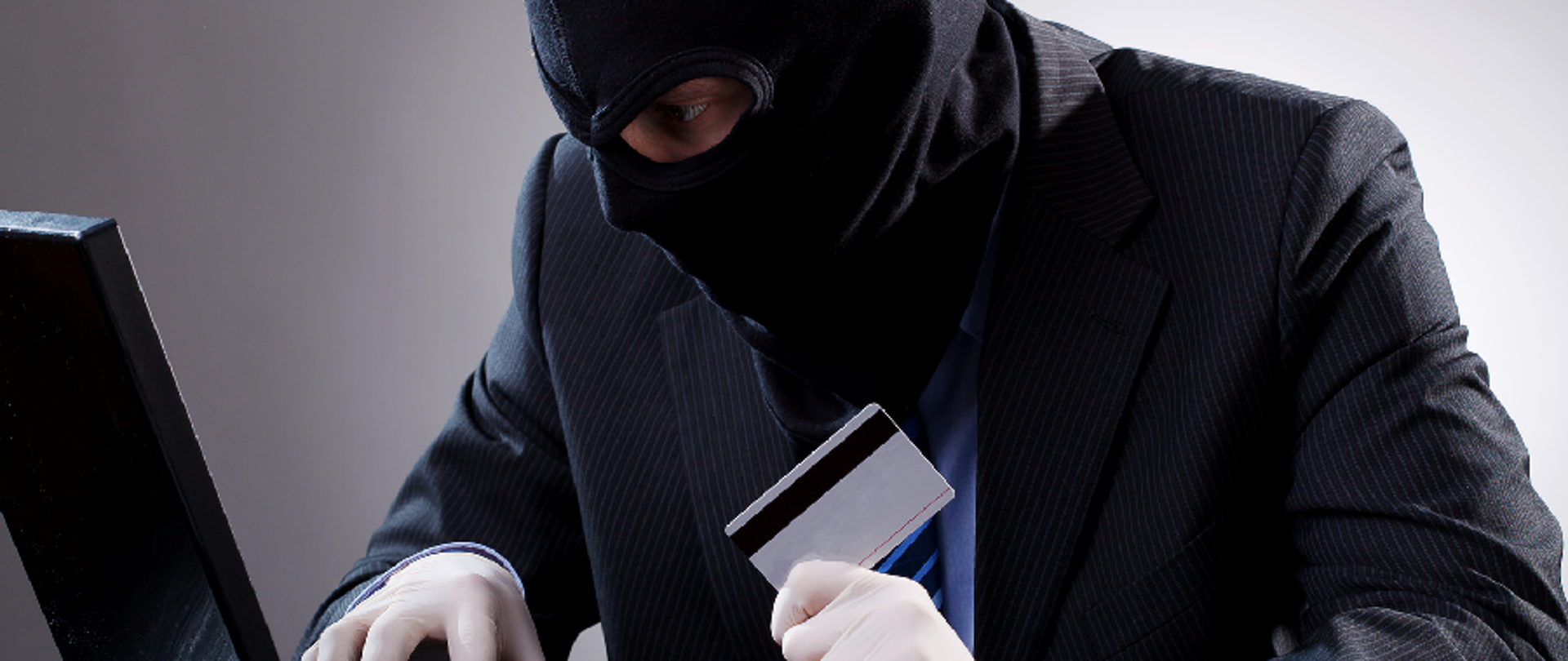 Mężczyzna w kominiarce i rękawiczkach, z kartą kredytową w dłoni, przy laptopie.