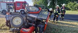 Ciągnik rolniczy lezy rozbity na poboczu drogi z jednej strony stoi osoba postronna natomiast z tyłu ciągnika trzech strażaków jeden nachyla się , za nimi na drodze samochód strażacki w oddali zielone drzewa