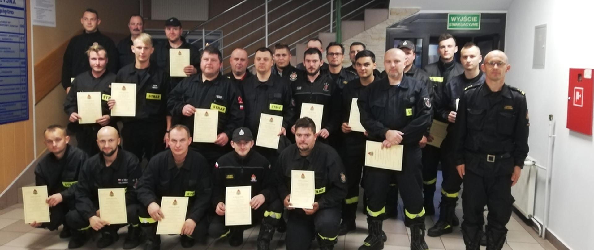 Na zdjęciu widoczni strażacy na holu komendy Powiatowej PSP w Gorlicach w ubraniach koszarowych koloru czarnego bez nakrycia głowy trzymający w rękach dyplomy ukończenia kursu. 